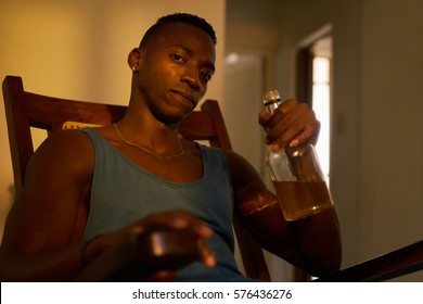 Social issues, substance abuse with alcoholic young black man drinking alchool from liquor bottle at home, looking at camera holding bottle of whiskey.