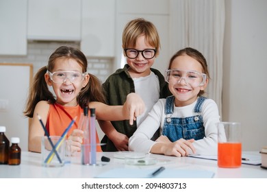 Social Little Kids Doing Home Science Project, Sharing Their Fun Experience. All Behid Table, Wearing Glasses. Chemical Glassware And Colored Liquids On The Table.