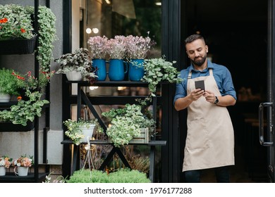 Social Media, Ad And Online Offer For Small Business Owner. Smiling Bearded Millennial European Male In Apron Watch On Smartphone At Front Door Of Flowers Rustic Shop With Different Plants, Free Space