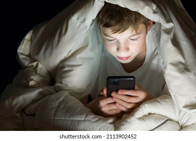 Social Media Addiction Boy Hiding Under The Blanket At Night In His Bed Communicates On Internet. Child Gadget Addiction And Insomnia. Lonely Boy Using Smartphone At Night In Bed