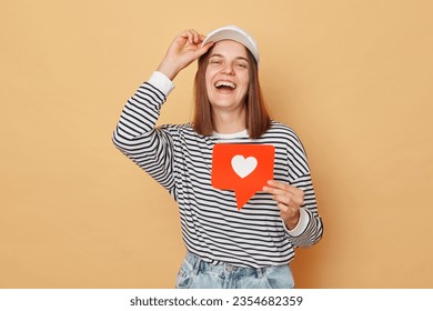 Social media content sharing. Notification for recommended content. Counting likes and clicks. woman wearing shirt and baseball cap holding like blogger icon standing isolated  over beige background