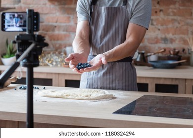 Social Media Influencer Or Food Blogger Creating Content. Man Shooting A Cooking Video Using Camera On Tripod. Chef Holding Bluberries In Hands And Showing Them To Viewer.