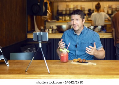 Social media influencer or food blogger creating content inside small restaurant – man sharing online food review using smartphone on tripod and lavalier, smiling content creator vlogger filming video