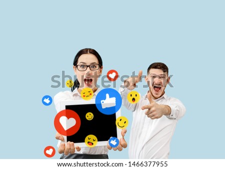 Social media interactions on mobile phone. Internet digital marketing, Chating, commenting, liking. Smiles and icons above tablet screen, that holding by young couple on blue studio background.