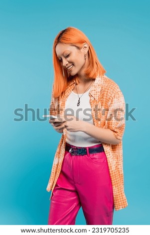 social networking, cheerful asian woman with dyed hair messaging, using smartphone, standing on blue background, smiling, orange shirt, casual attire, digital native, generation z