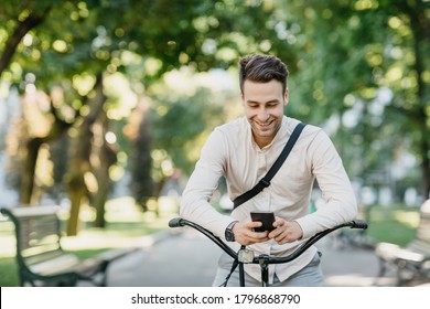 Social networks outdoors. Smiling attractive guy in shirt and trousers sits on bicycle in park and looks at smartphone, free space