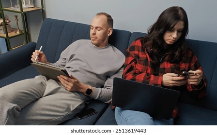 Social trend. Young couple sharing time together with gadgets sitting on sofa in home interior. Digital activity, remote works or modern emotional disconnect concepts - Powered by Shutterstock