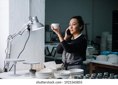 Social Woman Examening A Cup Under A Lamp On A Table With Newly Produced Blank Tableware And Tools. In A Pottery Workshop. Making Report On The Phone.