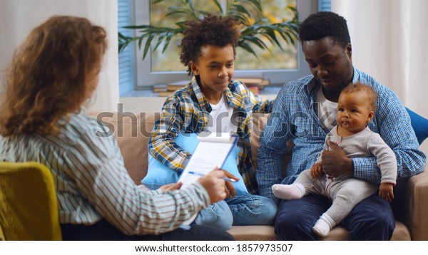 Social worker consulting\
smiling young single father with kids at home. Case worker woman\
visiting african man with schoolboy son and baby daughter providing\
consultation