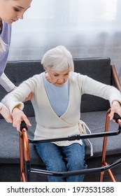 Social Worker Helping Elderly Woman Getting Up From Sofa Near Medical Walkers