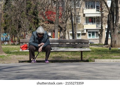Sofia, Bulgaria - April 01, 2021: Retired woman with white hair, sunglasses and cane checks what time it is while sitting on a park bench