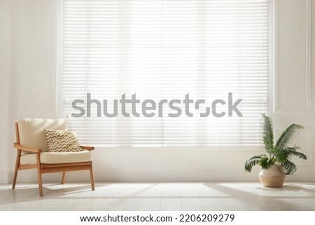 Soft armchair and houseplant near large window with blinds in spacious room. Interior design