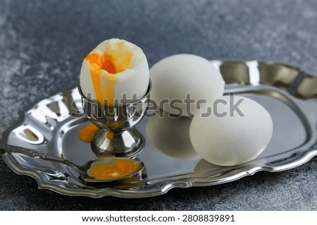Soft boiled eggs sit in a silver egg cup with a spoon nearby on a shiny silver tray. Two unpeeled eggs rest next to the cup. This scene shows a breakfast setting.