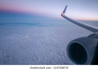 Soft Cloudscape In Predawn Glow From Airplane Window With The Wing And Jet Engine On The Right. First Blush Of Morning After An Overnight Red Eye Flight