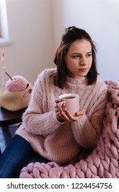 Soft Cozy Photo Of Slim Girl In Warm Sweater With Cup Of Tea In Hands. Girl Sitting On Giant Knitted Plaid. Knitting, Wool Thread Needles. Young Woman At Home Sitting On Chair Relaxing, Drinking Tea