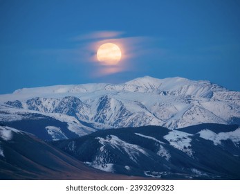 Soft focus. Big moon over a snowy mountain. Night replaces the full moon day. Golden full moon rising over distant mountains in the blue hour. - Powered by Shutterstock