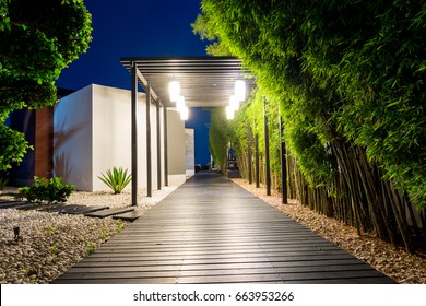 Soft Focus Of Black Wooden Walk Way With Bamboo On Both Side Over White Pebbles Floor At Night, Tropical Garden Path Decorative Lighting By Bulb Hung On Facade In Warm White Light.