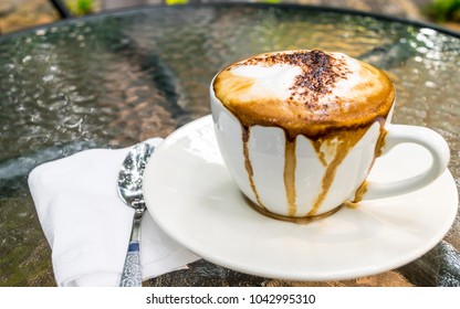 Soft Focus And Blurred Of A White Cup Of Hot Latte Coffee, Mess Up, With Green Leaf Background Texture, Overflow On Cup, On Glass Table