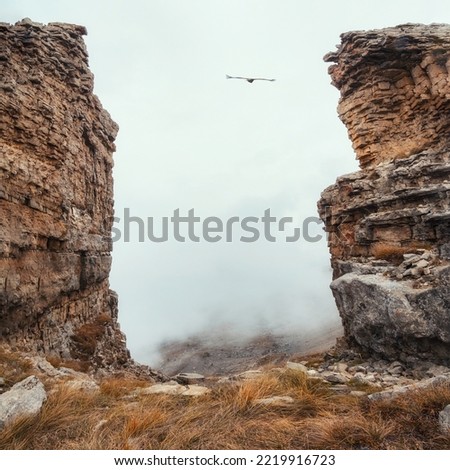 Soft focus. Dangerous gorge, a cliff between two rocks. Dramatic fog among giants rocky mountains. Ghostly atmospheric view to big cliff in cloudy sky. Low clouds and beautiful rockies. 