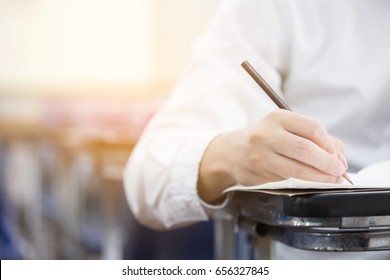 Soft Focus Front View Undergraduate Holding Pencil Writing On Paper Answer Sheet And Sitting On Lecture Chair Doing Final Exam Attending In Examination Room Or Classroom.university Student In Uniform.