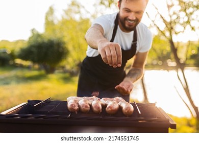 Soft focus of happy male chef in apron smiling and adding salt to sausages and meat on grill, while preparing food for picnic on summer day in countryside - Powered by Shutterstock