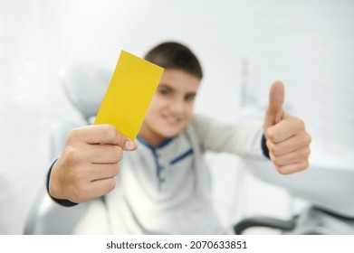 Soft Focus On Blank Yellow Visit Card With Copy Space For Insert Ads And Text In The Hand Of Blurred Handsome Boy Smiling Toothy Smile And Showing Thumb Up Looking At Camera Sitting In Dentist Chair