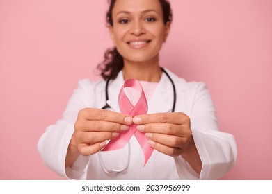 Soft focus on Breast Cancer Awareness Pink Ribbon, in hands of blurred mixed race woman doctor in white medical coat, isolated on colored background. 1 st October, World Day of fight breast cancer