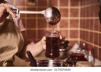 Soft focus on a ladle and a glass jar in the hands of a housewife, a confectioner pouring freshly made cherry jam into a sterile jar in a home kitchen. Woman preparing homemade canned food