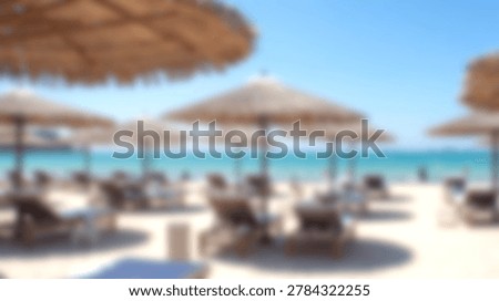 A soft focus view of multiple straw beach umbrellas and lounge chairs arranged on a sandy beach overlooking the turquoise ocean water