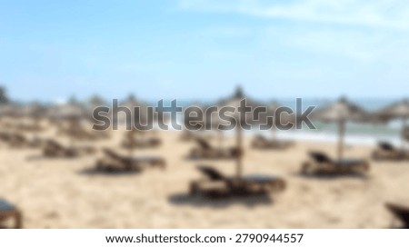 A soft focus view of multiple straw umbrellas and wooden lounge chairs arranged on a sandy beach shoreline under a bright sky