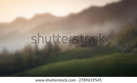 A soft focus view of a rustic cabin situated on a green slope with dense fog rolling over distant mountain peaks in the early morning light