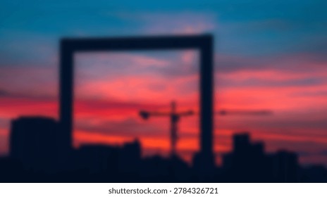 A soft focus view of a tall rectangular building frame and a construction crane silhouetted against a colorful sunset sky in an urban environment