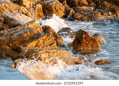 The soft glow of sunset casts light over the waves, which crash onto the rocks, sending splashes into the air, while the ocean reflects golden hues, offering a calming and natural coastal view.