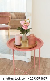 Soft home decor in a gentle tonality, a pink jug, a candle, a vase with pink beautiful flowers on a white wall background and in a round pink table a tray. Interior.