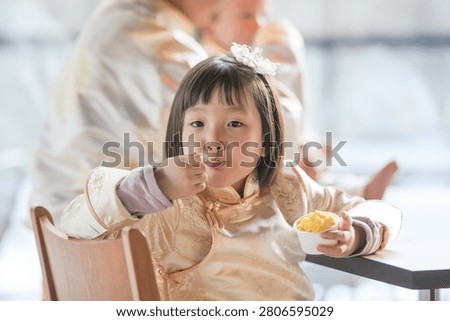 Soft light shines on a smiling child. She eats a small bowl of food with joy. Her hands hold the bowl and spoon. Her hair is adorned with a small white bow. Her eyes sparkle with delight