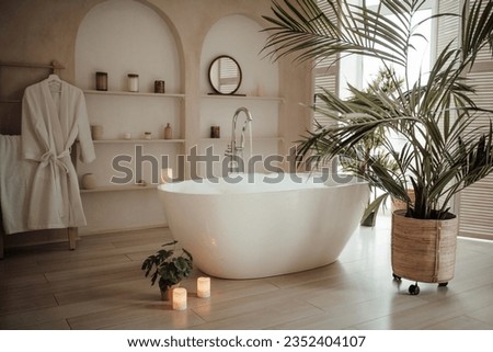 Soft native hues organic shapes look of bathroom with big window oval bathtub in neutrals tones. Green palm plants candles bubblebath leasure and relaxation skin selfcare wellness luxury living
