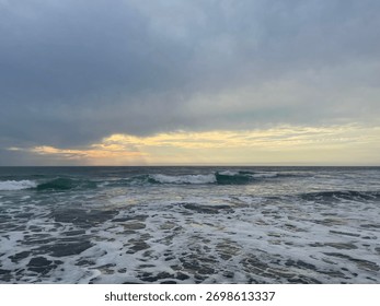 Soft Ocean Waves Rolling Toward the Shore Under a Cloudy Sky at Sunset, Gentle Light Reflects on the Water Creating a Peaceful Coastal Evening Scene