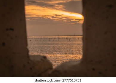 Soft orange sunset seen through aged metal edges, overlooking shallow waters lined with fishing poles. A layered composition blending texture, light, and coastal livelihood in a tranquil atmosphere.