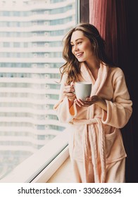 Soft Photo Of Fresh Young Woman In Pink Tender Bathrobe Drink Tea And Looking At Window . Pretty Girl With Perfect  Wavy Hairs Enjoying  Early  Sunny Morning.