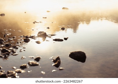 Soft smooth haze in golden sunbeams on water surface of calm river with ray, blinks, glares, reflection and stone on shore, silent and quiet wild nature background, texture, detail. - Powered by Shutterstock