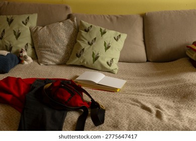 Soft sofa strewn with jacket and backpack. Open notebook on throw, green patterned cushions, warm yellow wall, lateafternoon light, livedin texture and casual domestic clutter 