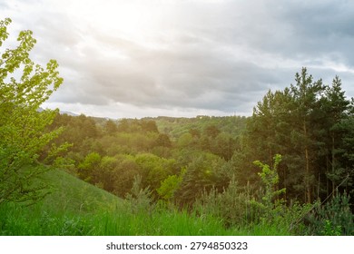 Soft sun rays breaking through storm clouds over dense forest valley. Mountainous summer landscape with green hills and mix of deciduous and pine trees.