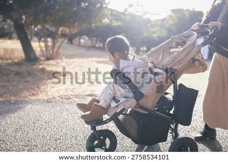 Soft sunlight glows behind trees. Child rests in stroller, eyes closed, calm. Parent walks beside, gentle presence. Scene captures quiet, peaceful outing. Ideal for parenting, family