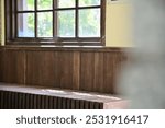 Soft sunlight passes through the window of a wooden Japanese-style train station, casting gentle shadows on the interior chairs. The light highlights the outdoor greenery, creating a peaceful ambiance