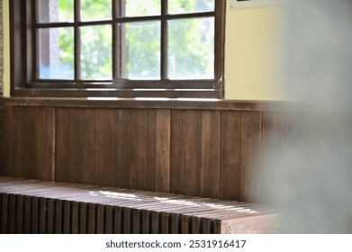 Soft sunlight passes through the window of a wooden Japanese-style train station, casting gentle shadows on the interior chairs. The light highlights the outdoor greenery, creating a peaceful ambiance