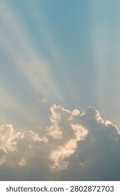Soft sunrays streaming through white and golden cumulus clouds against a pale blue summer sky