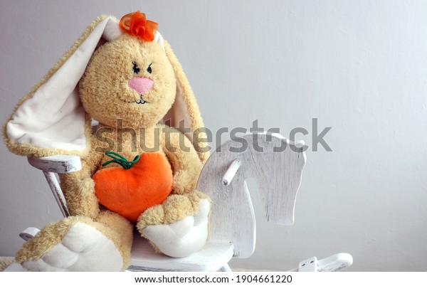 Soft toy-yellow hare with\
a heart in his hands on a wooden horse on a gray background, space\
for text