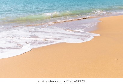 Soft waves gently lap against a sandy beach, creating a foamy edge where water meets shore. The clear blue sky reflects on the tranquil water, enhancing the serene coastal scene