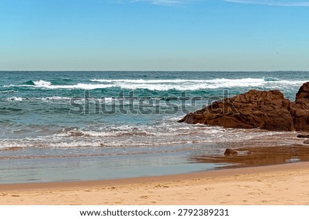Soft blue‑green waves roll onto a smooth sandy beach while a rugged rocky formation juts into the water on the right, catching the incoming swell. Whitecaps shimmer across the surface beneath a clear light‑blue sky, creating a calm coastal scene defined by texture contrast, natural colour, and quiet seaside movement.