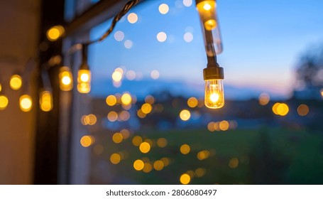 Softly glowing string lights create a bokeh effect against a twilight sky, evoking a cozy and festive atmosphere.